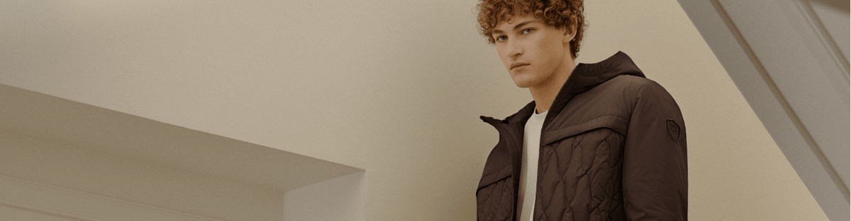 A man wearing a dark brown quilted hooded jacket, standing in a minimalist modern interior with beige walls and white structural beams.