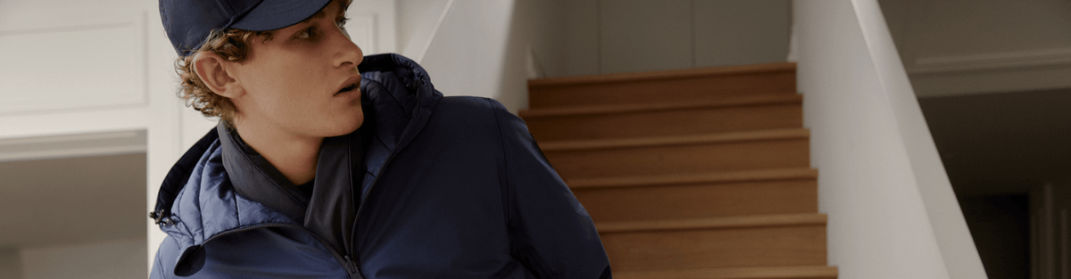 A person wearing a navy blue jacket and matching cap standing indoors near wooden stairs.