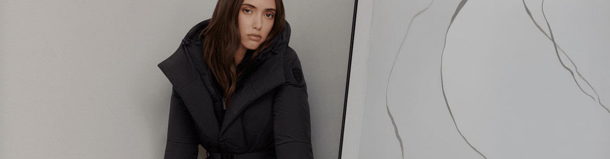 A woman wearing a black hooded puffer jacket with a belted waist, posed against a minimalist light gray wall with a large abstract art piece on the right.