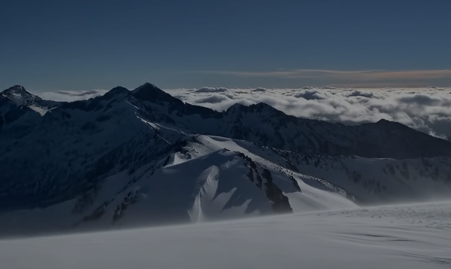 Snowy mountain landscape with peaks and valleys, partially covered by clouds.